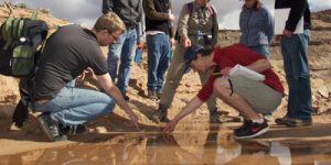 two students crouch in shallow water to gather samples. Six others stand behind them looking on. Desert hills and a blue sky with fluffy clouds in background"