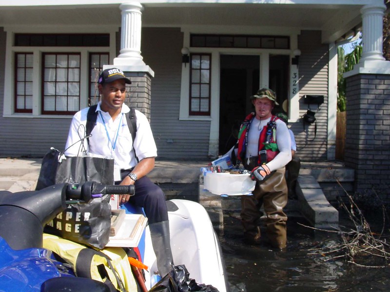 Rescuer on a water craft in front of a flooded home where a man wearing waders is carrying items out of the house.