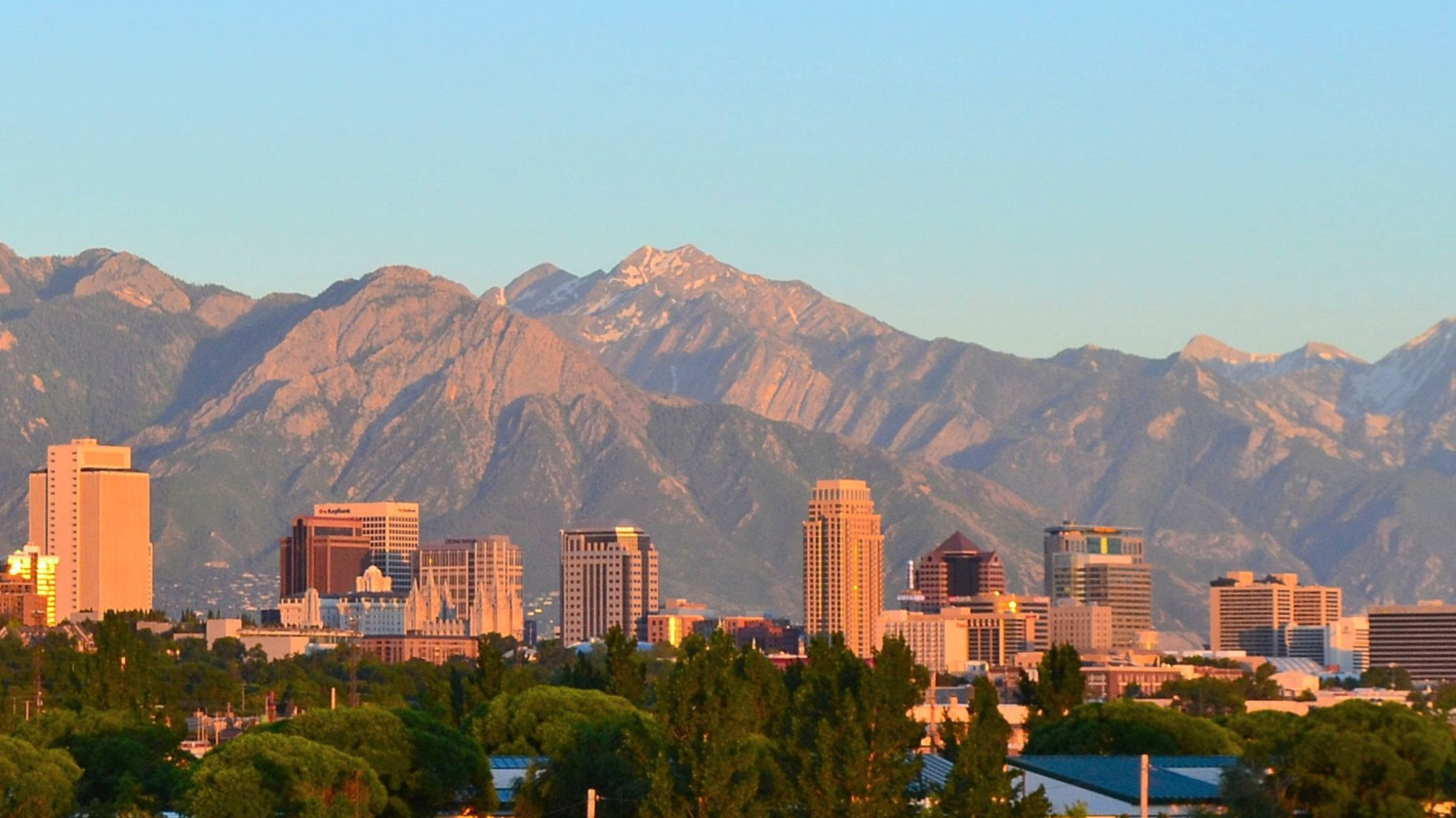 Tall buildings in Salt Lake City against the Wasatch Mountains, under a blue sky