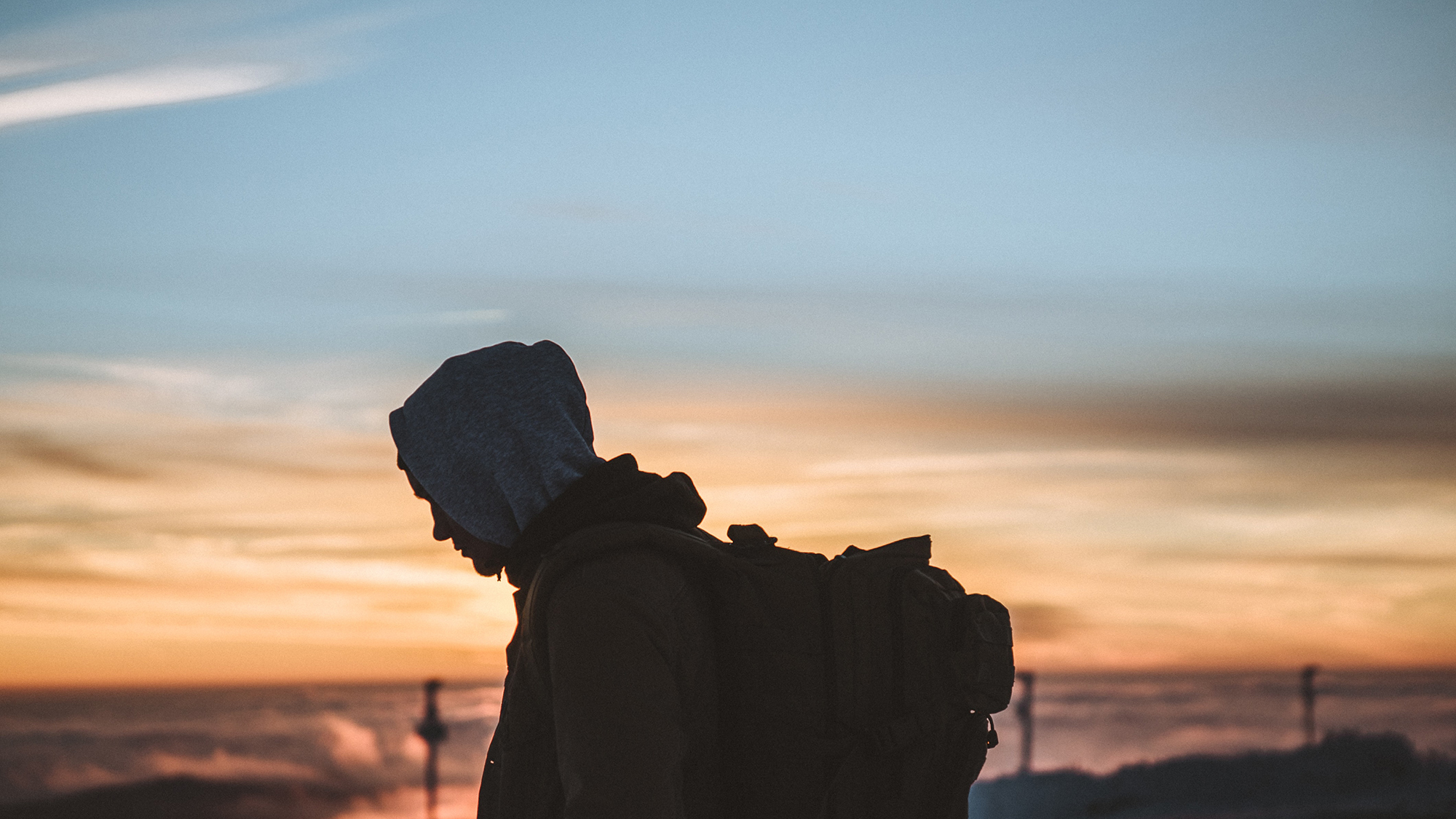 profile of a person wearing a hoodie and a large backpack against an orange sunset