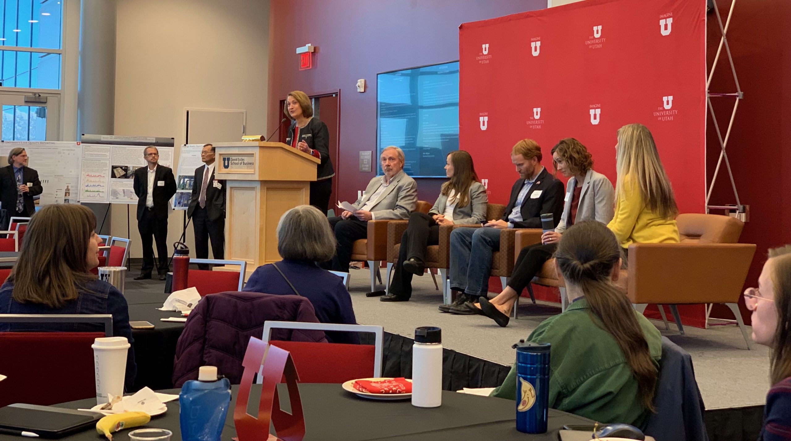 President Ruth Watkins stands at a podium next to five seated panelists. Attendees are seated and standing around the room.