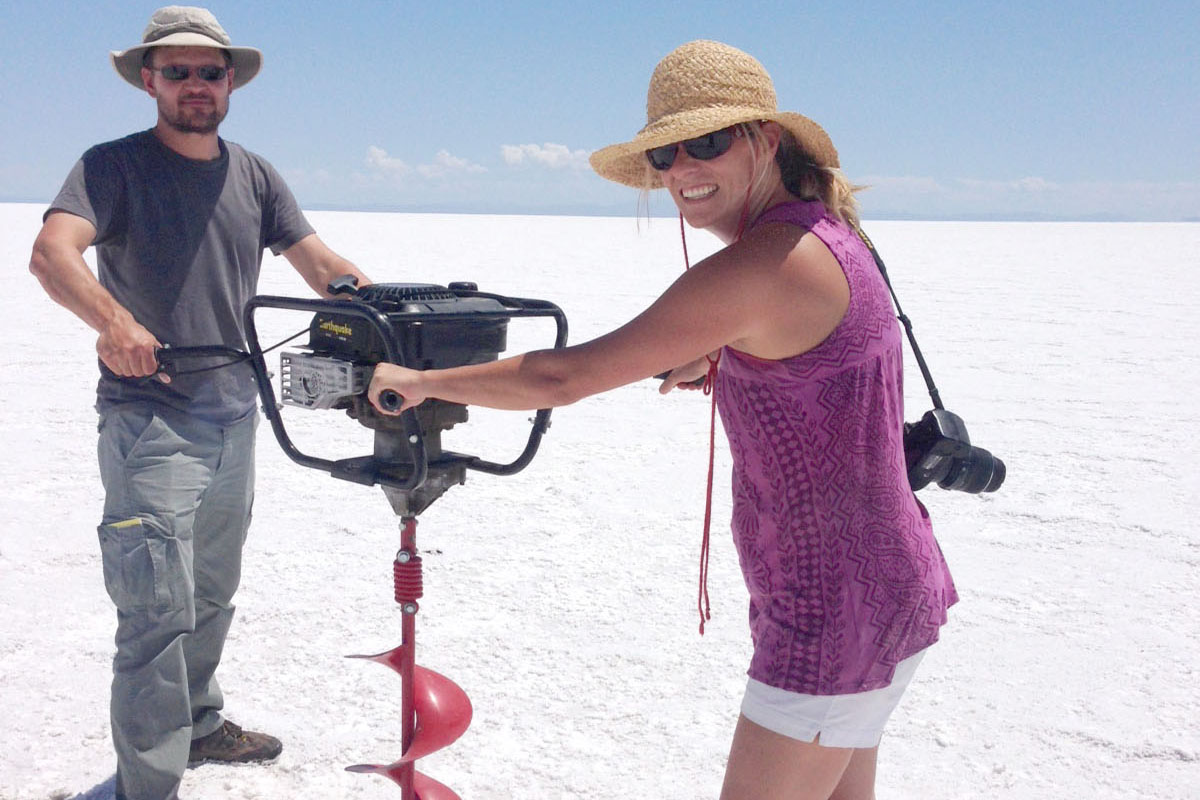 A man and a woman are standing on a white expanse of salt under a blue sky. They are gripping the four handles of a chest-high industrial drill.