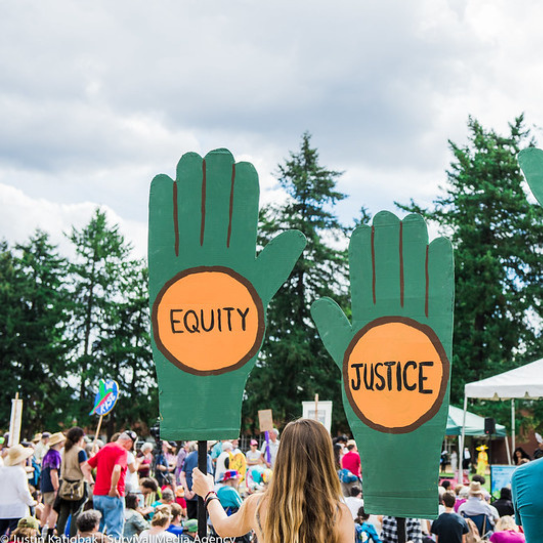 At the edge of a large colorfully dressed crowd of people, a woman is holding aloft two large green signs in the shape of hands, with words in orange circles: equity on the left sign and justice on the right.